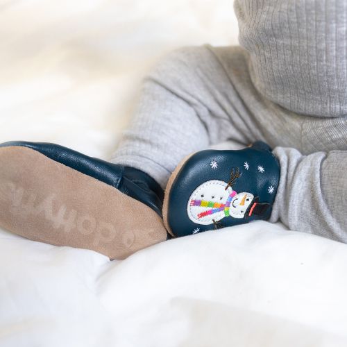 Baby boy sitting, wearing navy Dotty Fish barefoot shoes with white snowman wearing black hat and embroidered rainbow scarf design.