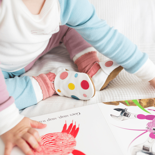 Toddler reading a book, wearing white leather Dotty Fish first walker shoes with pink ankle trim and multicoloured spots.