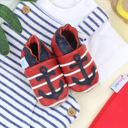 Red leather baby shoes with navy anchor, sitting with baby clothing.